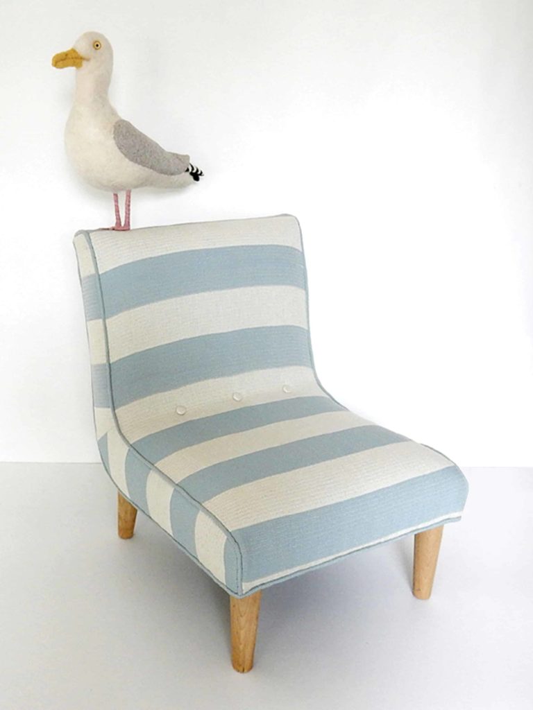 Needle felted seagull with bag of chips, standing on a little chair upholstered in blue and white striped fabric.