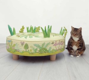 Cat sitting beside a child's round play stool; fabric with cute animals, bugs, birds, and flowers. Grass and leaves stitched on top.