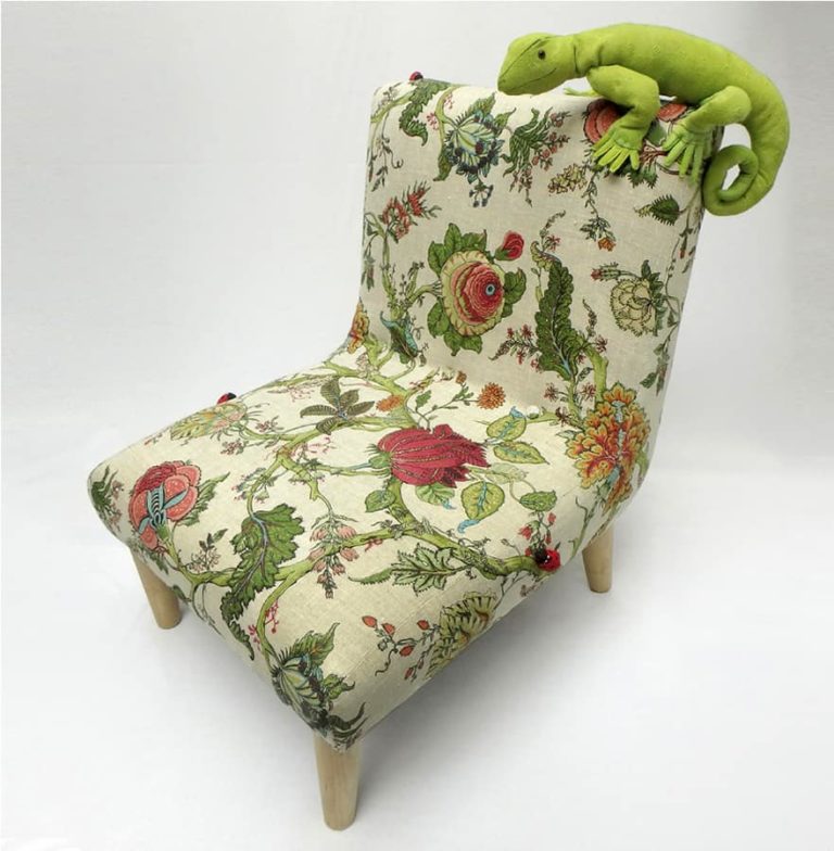 Cute, green fabric lizard sitting on a little chair, upholstered in floral botanical linen fabric, surrounded by real plants.