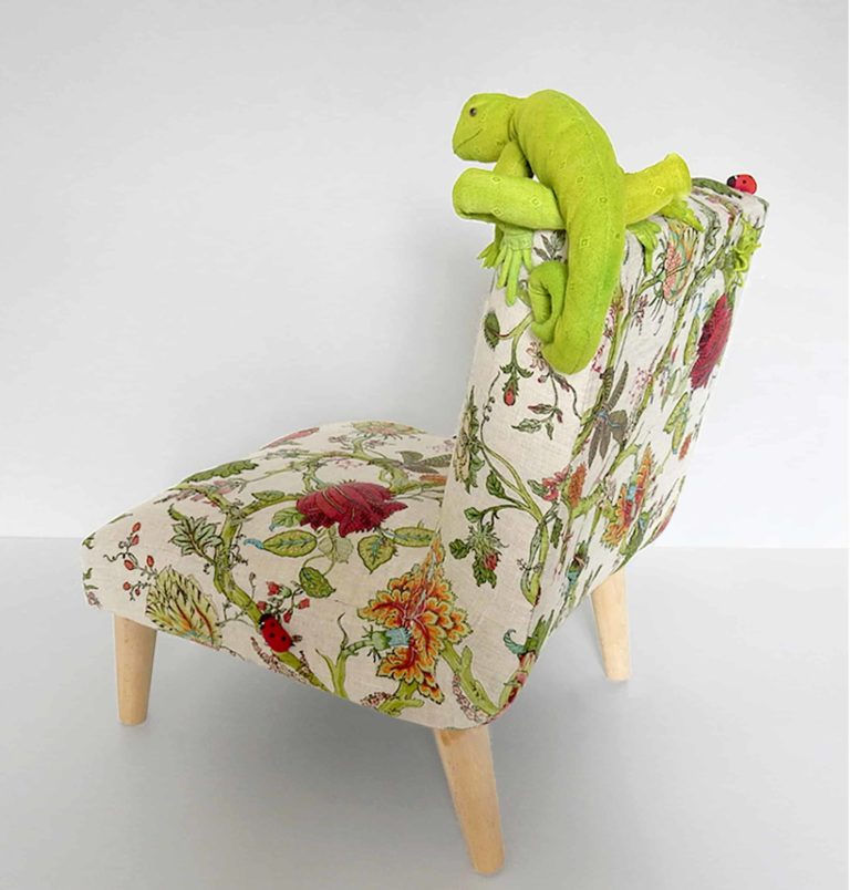 Cute, green fabric lizard sitting on a little chair, upholstered in floral botanical linen fabric, surrounded by real plants - rear view
