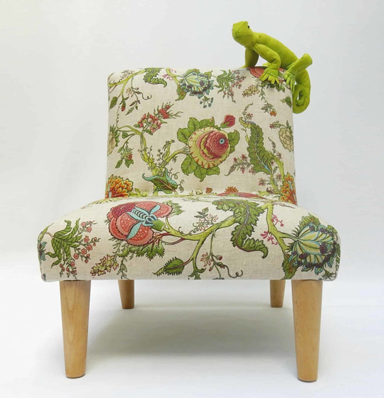 Cute, green fabric lizard sitting on a little chair, upholstered in floral botanical linen fabric, surrounded by real plants. Front View..
