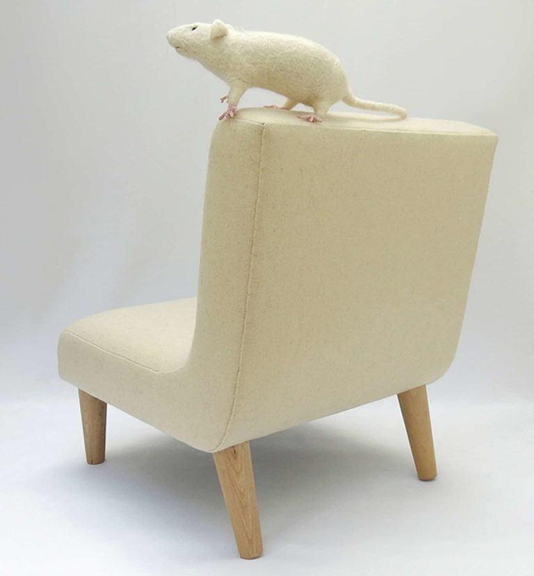 Needle felted white rat sitting on top of modern child's chair in pale cream wool fabric. Rear view.