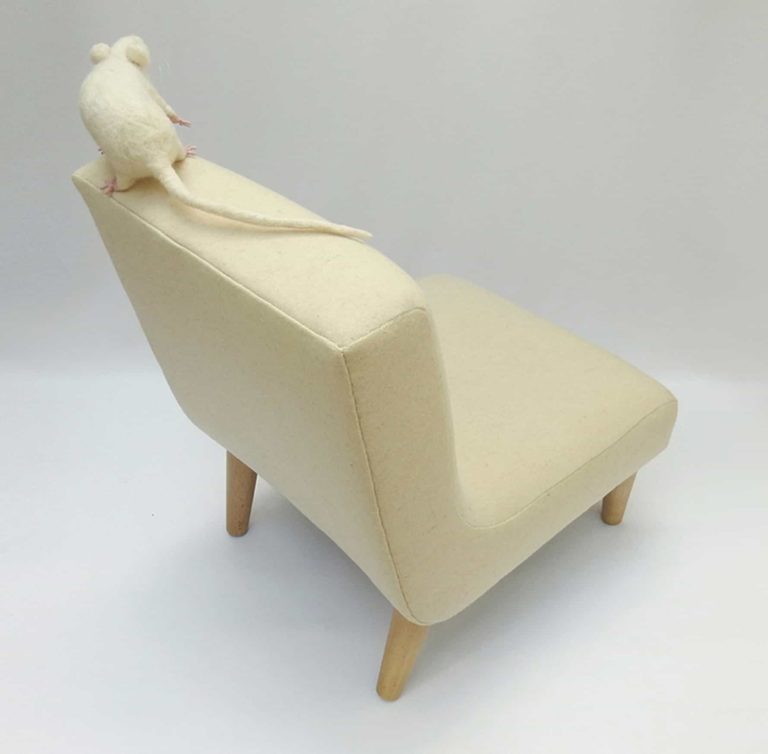 Needle felted white rat sitting on top of modern child's chair in pale cream wool fabric. Top view.