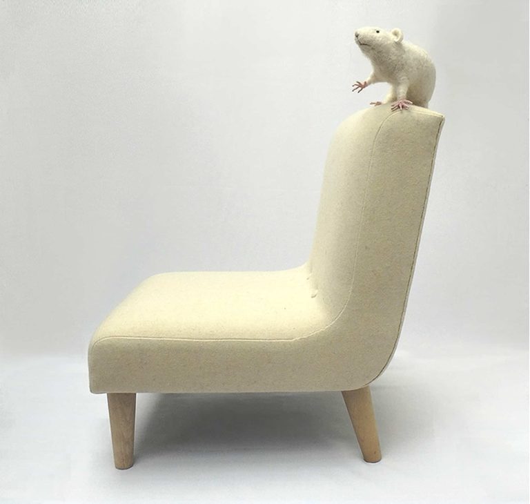 Needle felted white rat sitting on top of modern child's chair in pale cream wool fabric. Side view.