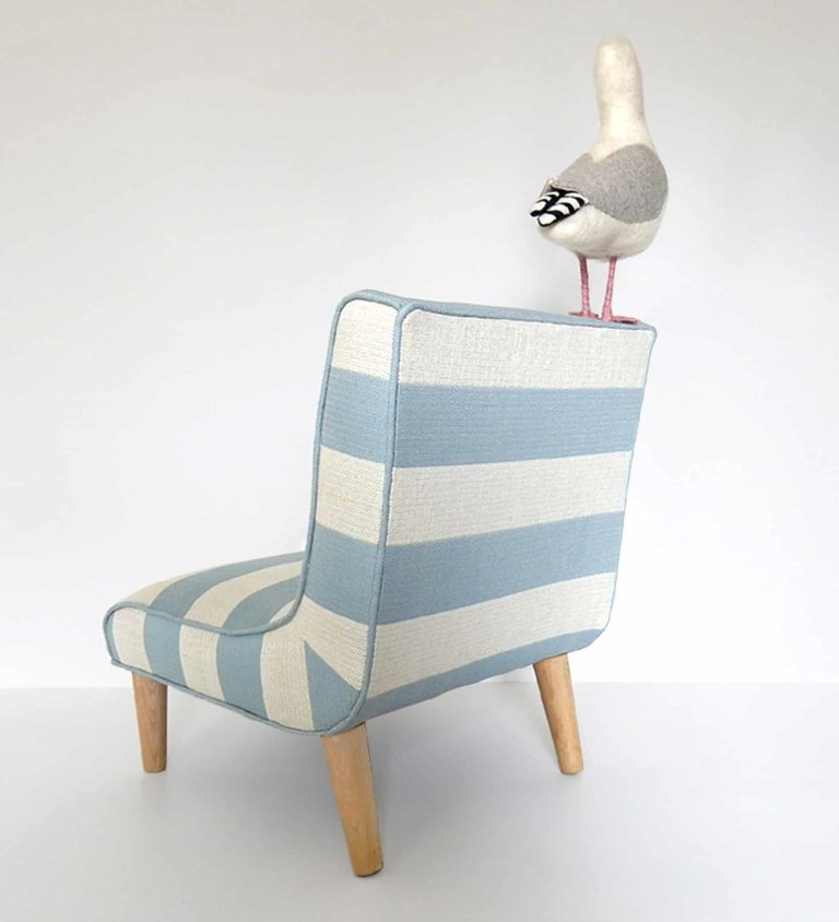 Rear view of needle felted seagull with bag of chips, standing on a little chair upholstered in blue and white striped fabric.