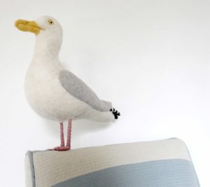 Close-up of needle felted seagull with bag of chips, standing on a little chair upholstered in blue and white striped fabric.