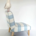 Needle felted seagull with bag of chips, standing on a little chair upholstered in blue and white striped fabric - side view
