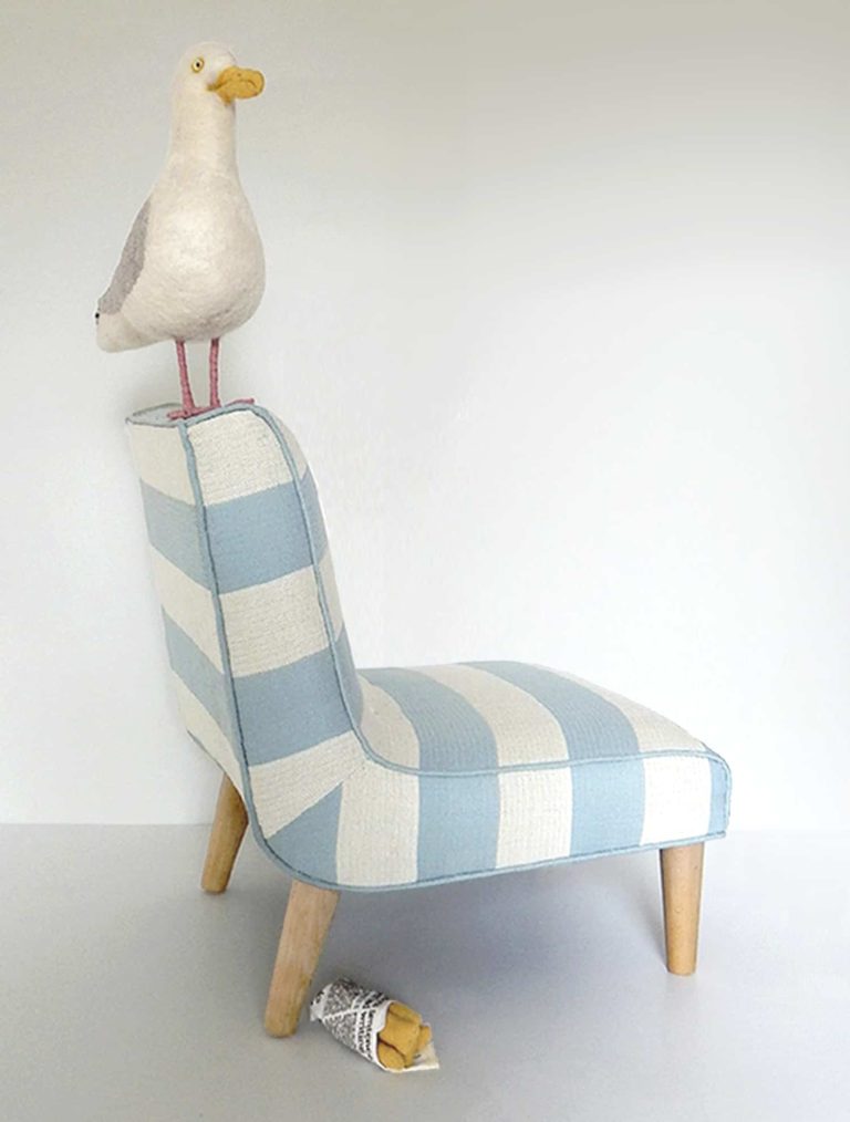 Needle felted seagull with bag of chips, standing on a little chair upholstered in blue and white striped fabric - side view