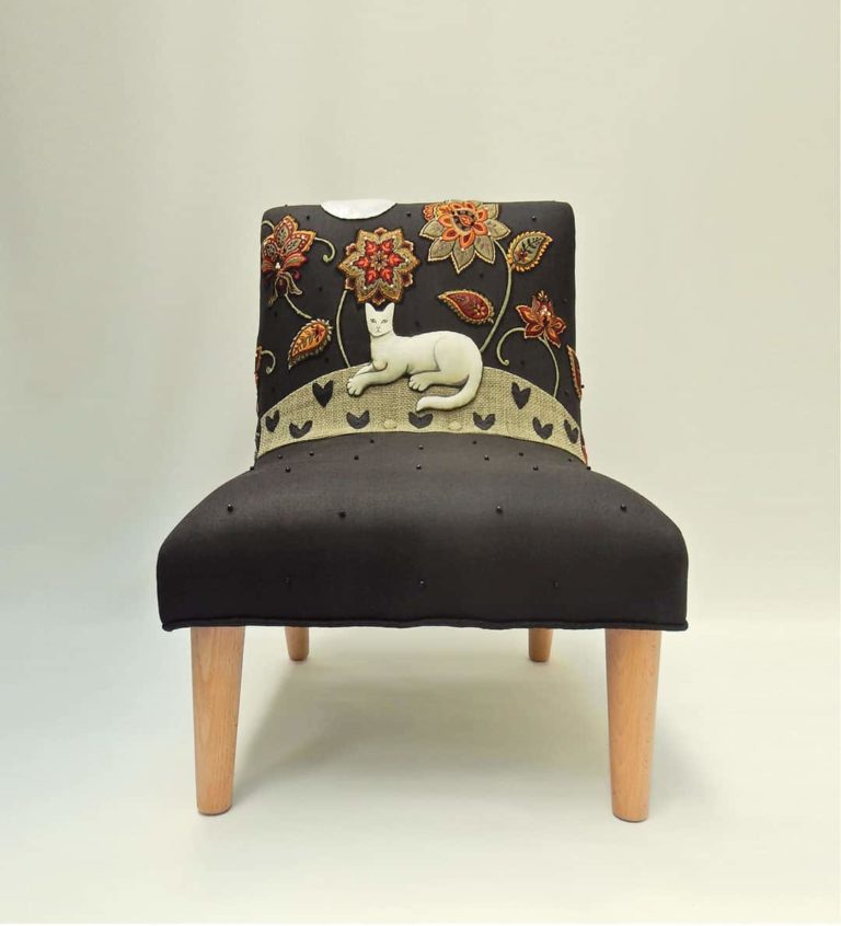 Child’s chair, hand sewn appliqué fabric of a white cat lying down under red and gold embroidered flowers on black linen.