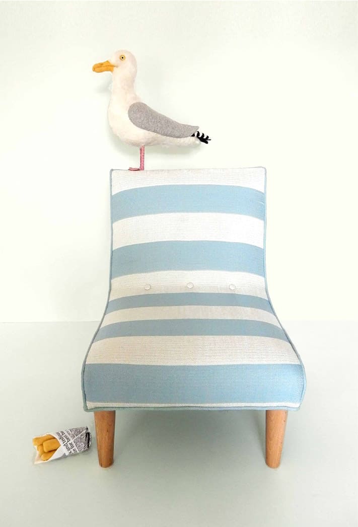 Needle felted seagull standing on a little chair, upholstered in blue and white striped fabric.